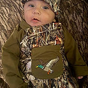 Archer joined the competition — help win amazing prizes! baby, child, infant, portrait, sitting, couch, cushion, camouflage, outfit, hat, onesie, bib, duck_patch, wide_eyed, face, indoors, textile, patterned_clothing, cozy, fashion