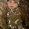 baby, child, infant, portrait, sitting, couch, cushion, camouflage, outfit, hat, onesie, bib, duck_patch, wide_eyed, face, indoors, textile, patterned_clothing, cozy, fashion