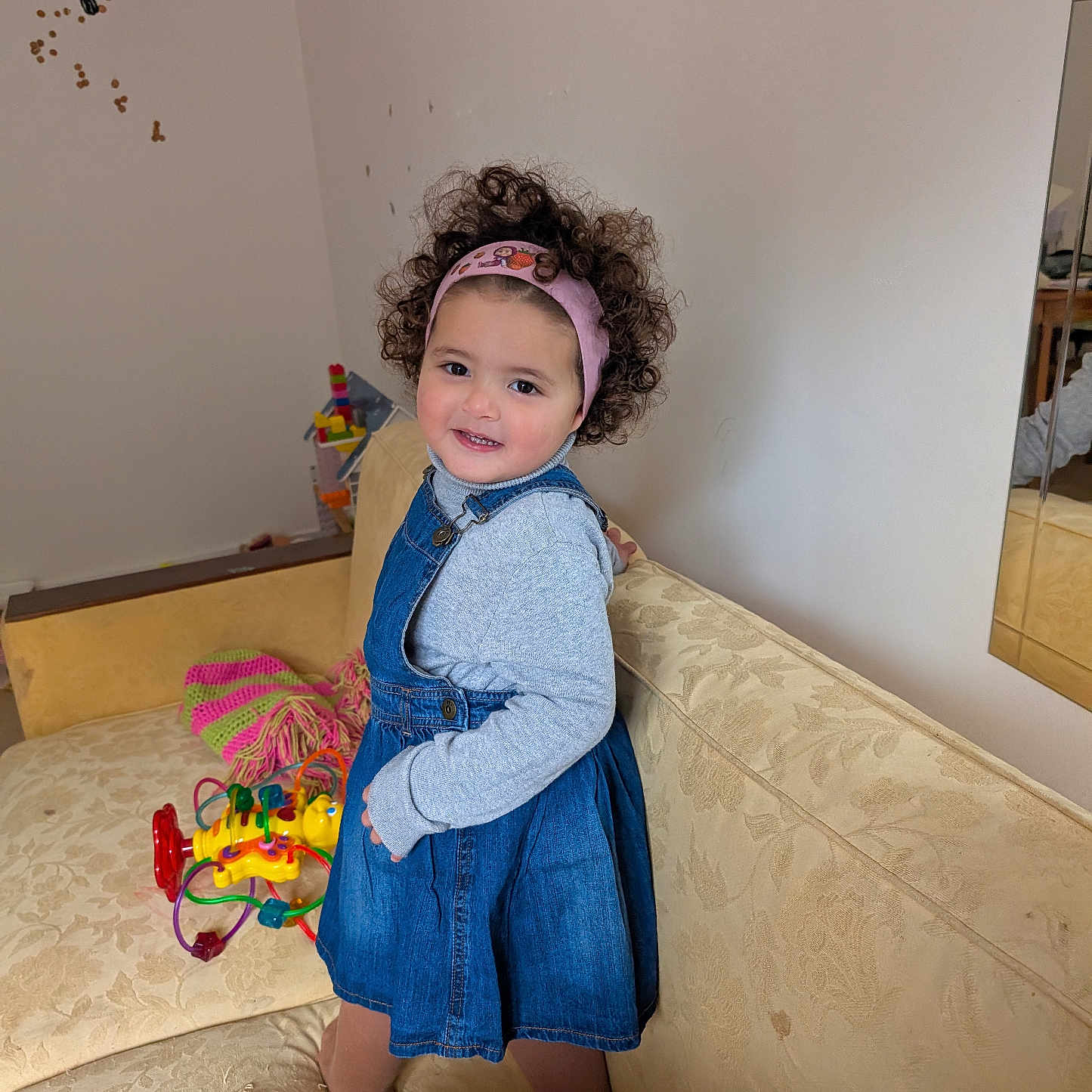 Nour-Elsa a rejoint le concours — aidez-le/la à gagner de superbes lots ! child, couch, curly_hair, cute, denim_dress, fabric, footwear, furniture, happy, headband, indoor, knitted_item, living_room, mirror, play, reflection, smile, toddler, toy, wall
