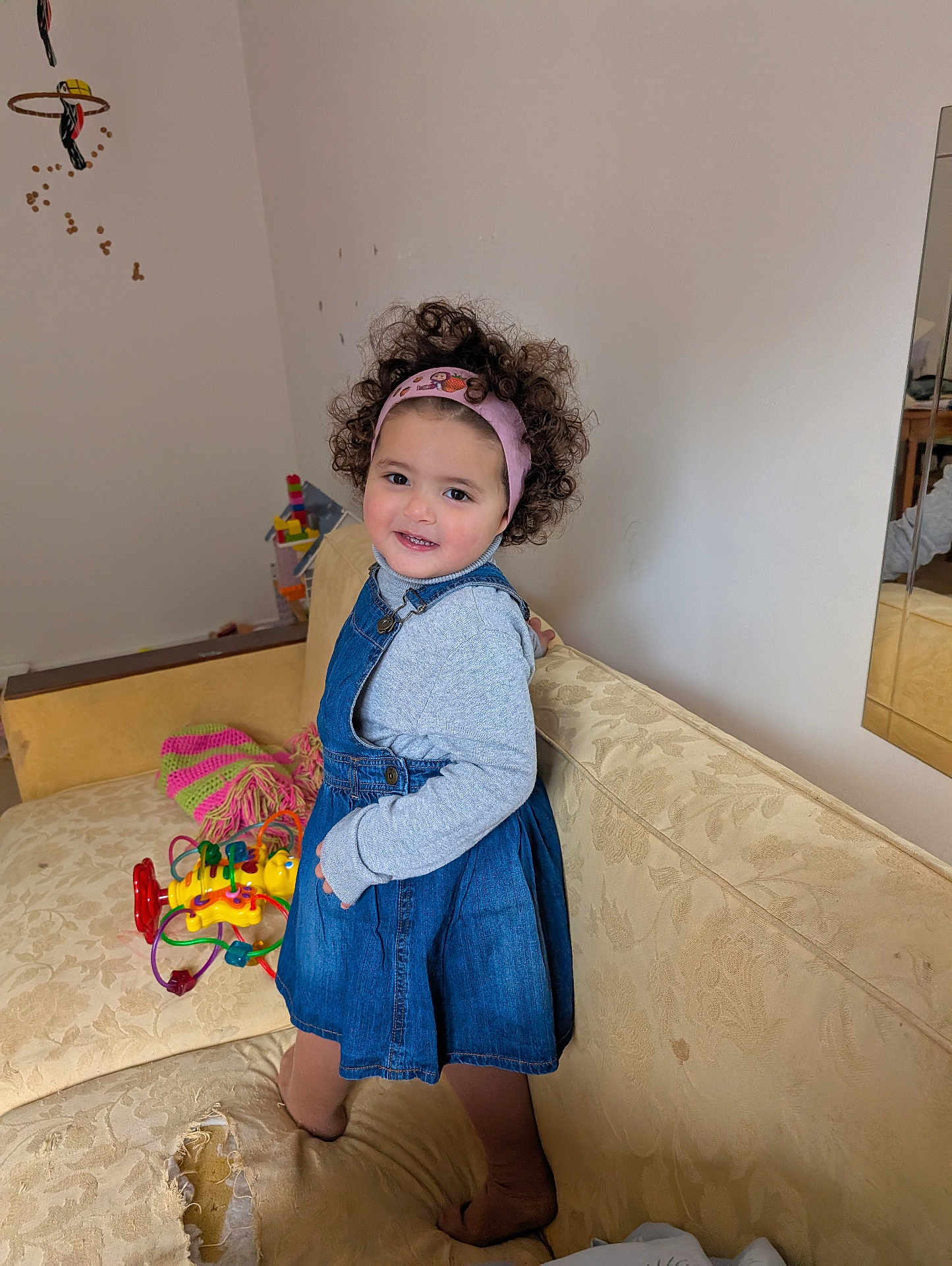 Nour-Elsa a rejoint le concours — aidez-le/la à gagner de superbes lots ! toddler, child, curly_hair, headband, denim_dress, couch, toy, knitted_item, smile, indoor, mirror, furniture, play, cute, happy, living_room, fabric, footwear, wall, reflection