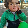 Nour-Elsa participe au concours pour gagner de l'argent avec cette photo : child, toddler, curly_hair, green_jacket, balloon, outdoor, playground, face, smile, rosy_cheeks, person, park, daylight, casual_clothing, young_child, hand_on_face, trees, play_equipment, people, background