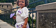 Nour-Elsa participe au concours pour gagner de l'argent avec cette photo : child, flower, sidewalk, dress, sandals, curly_hair, outdoor, fence, building, hill, sky, clouds, nature, smile, person, purple, white, urban, portrait, daylight