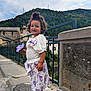 child, flower, sidewalk, dress, sandals, curly_hair, outdoor, fence, building, hill, sky, clouds, nature, smile, person, purple, white, urban, portrait, daylight