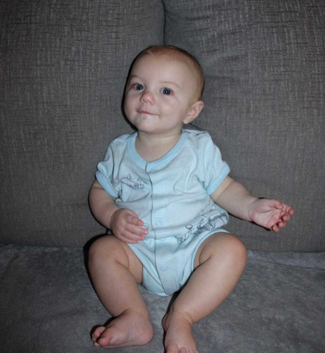 Titan is registered to the contest to win money with this photo: baby, child, infant, onesie, couch, gray, sitting, cute, face, skin, foot, hand, expression, young, indoors, smile, person, head, leg, blue