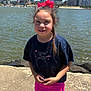 child, girl, waterfront, cityscape, skyline, sunny, outdoor, pink_bow, braid, shorts, tshirt, smile, concrete, rocks, water, buildings, daylight, casual_clothing, footwear, portrait