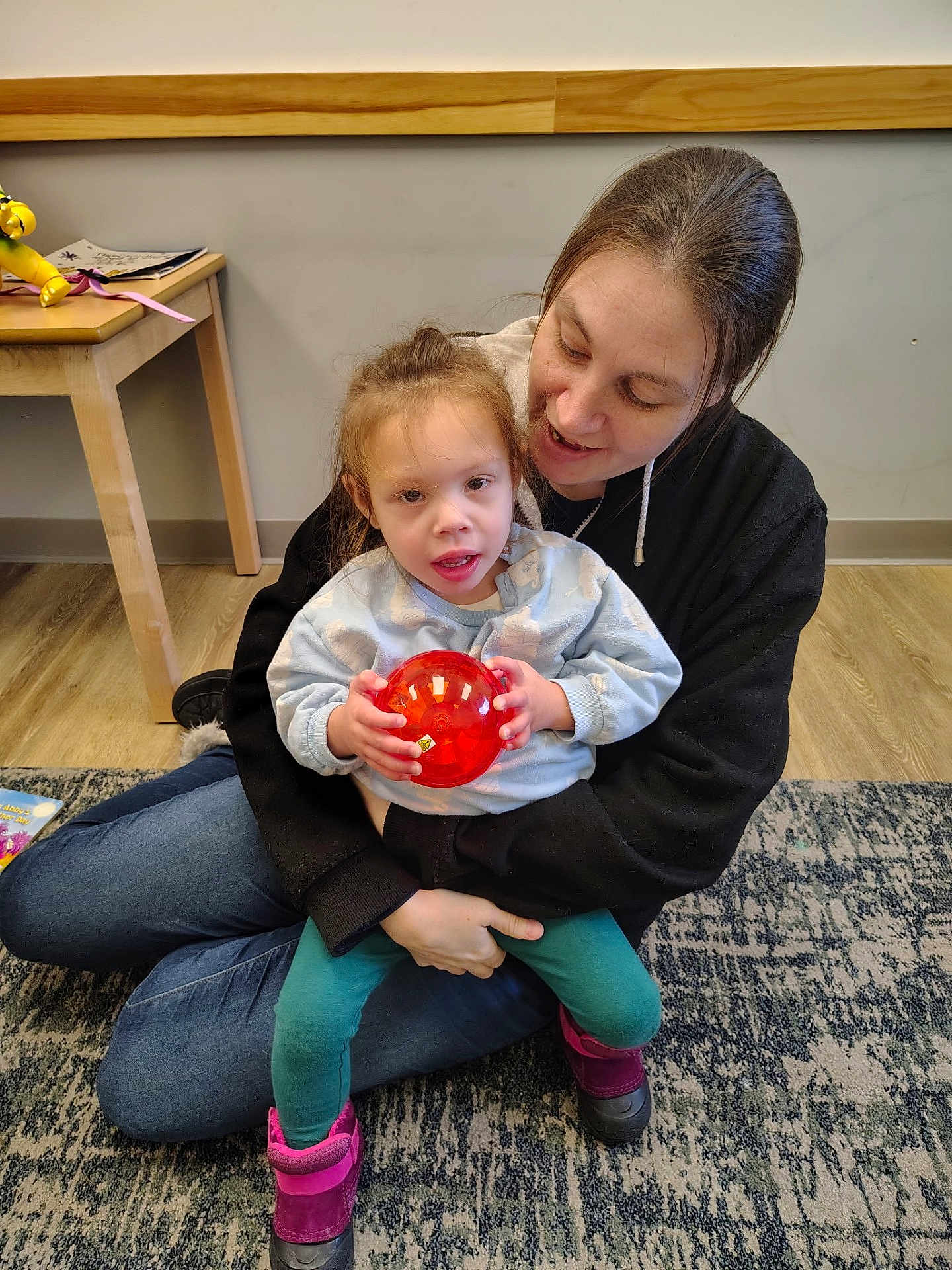 Lilith is registered to the contest to win money with this photo: child, woman, indoor, rug, toy, ball, floor, table, clothing, sitting, holding, smiling, person, wooden_furniture, casual, home, playtime, happy, young_child, cozy
