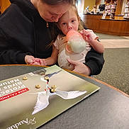 Lilith is registered to the contest to win money with this photo: child, adult, bottle, milk, table, calendar, cereal, indoor, library, person, holding, sitting, black_hoodie, light, wood, carpet, background, focus, intimate, comfort