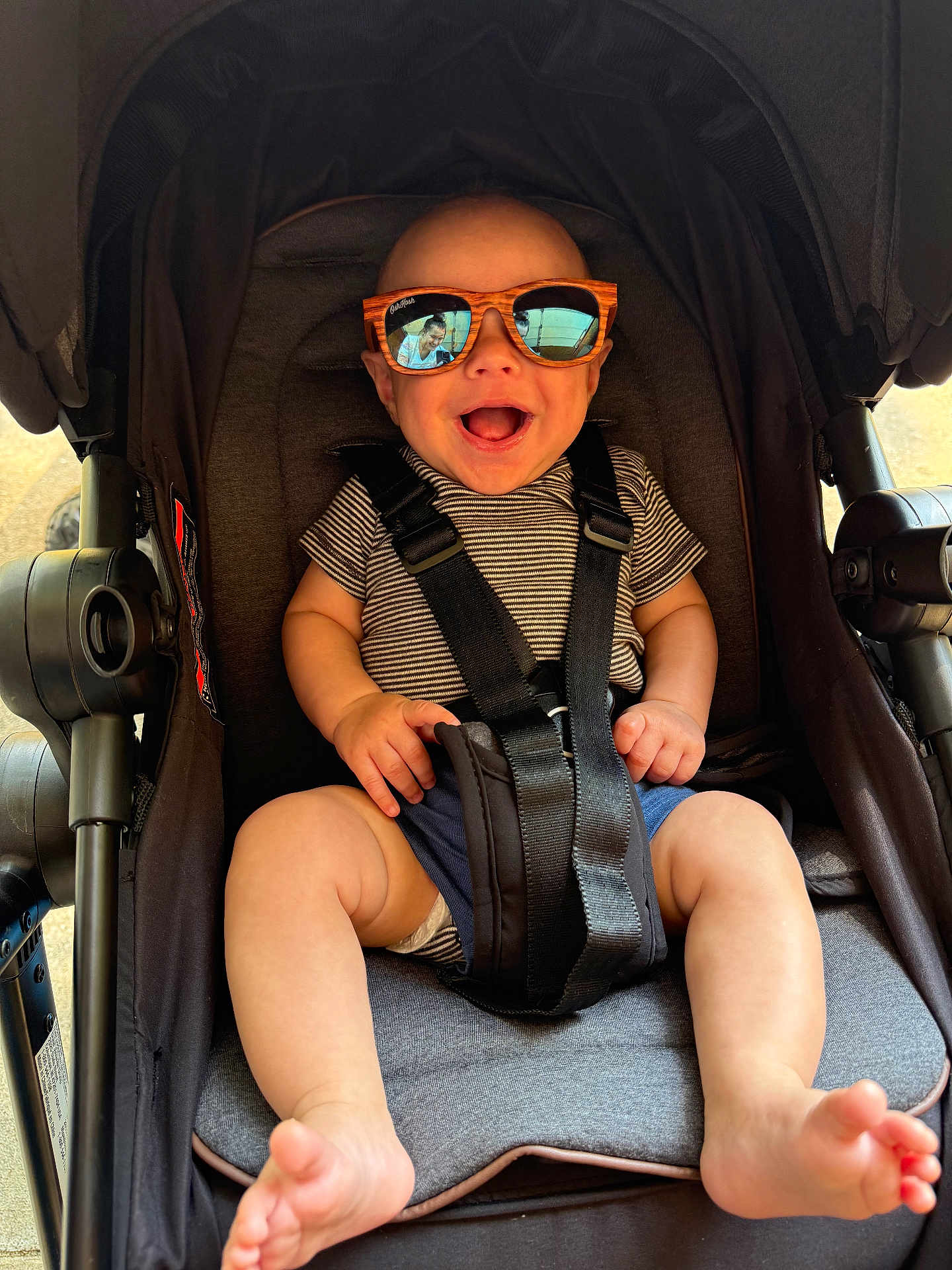 Colton is registered to the contest to win money with this photo: baby, barefoot, casual_clothing, child, closeup, comfort, happy, infant, legs, outdoor, person_reflection, reflection, seat, seatbelt, smiling, striped_shirt, stroller, sunglasses, sunlight, wooden_frame