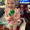 child, girl, toddler, smiling, happy, portrait, indoors, living_room, toy, green_toy, pink_shirt, long_sleeves, pants, socks, standing, face, blonde_hair, television, couch, blanket