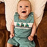 baby, smiling, green_checkered_romper, embroidered_tractors, couch, brown_couch, white_pillow, cozy, happy, child, infant, cute, indoors, portrait, sitting, clothing, hands, legs, face, blue_eyes