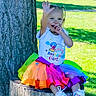 child, toddler, birthday, girl, smiling, waving, outdoor, park, tree, tree_stump, rainbow_tutu, colorful_clothing, birthday_shirt, sneakers, sitting, portrait, sunlight, grass, playful, happy