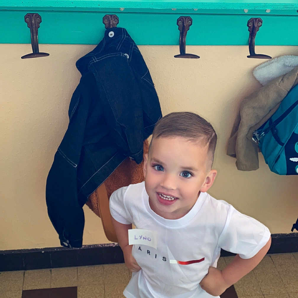 Lyno a rejoint le concours — aidez-le/la à gagner de superbes lots ! backpack, boy, casual_clothing, child, coat_hooks, hallway, happy, indoor, jackets, name_tags, person, playful, school, short_hair, smiling, sneakers, standing, tile_floor, white_tshirt, young_child