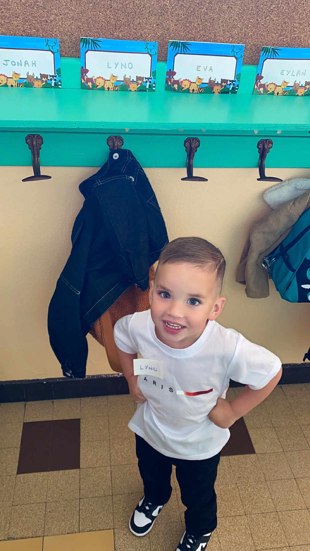Lyno a rejoint le concours — aidez-le/la à gagner de superbes lots ! backpack, boy, casual_clothing, child, coat_hooks, hallway, happy, indoor, jackets, name_tags, person, playful, school, short_hair, smiling, sneakers, standing, tile_floor, white_tshirt, young_child