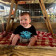 Ryett is registered to the contest to win money with this photo: baby, child, smiling, sitting, black_tshirt, gray_shorts, checkered_blanket, wooden_beams, wire_fence, indoor, farm, rustic, happy, person, face, head, blue_eyes, short_hair, natural_light, wood