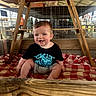 baby, child, smiling, sitting, black_tshirt, gray_shorts, checkered_blanket, wooden_beams, wire_fence, indoor, farm, rustic, happy, person, face, head, blue_eyes, short_hair, natural_light, wood