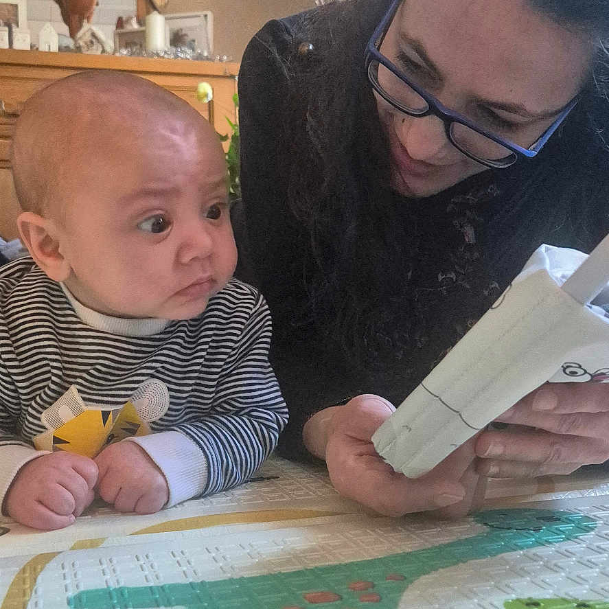 Louis a rejoint le concours — aidez-le/la à gagner de superbes lots ! baby, book, child, close_up, curious, curtains, expression, family, floor, glasses, home, indoor, interaction, person, play_mat, reading, striped_clothing, table, window, woman