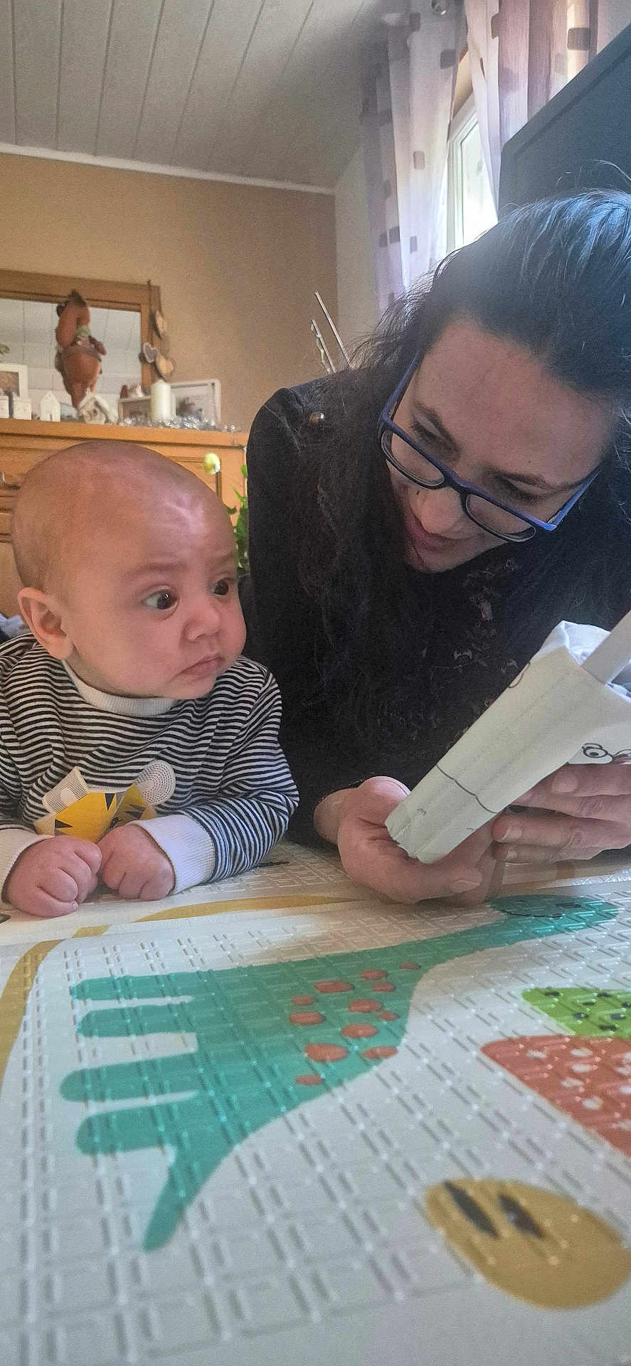 Louis a rejoint le concours — aidez-le/la à gagner de superbes lots ! baby, woman, glasses, reading, book, play_mat, indoor, curious, child, person, striped_clothing, floor, table, window, curtains, home, family, interaction, close_up, expression