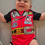 Lyzio a rejoint le concours — aidez-le/la à gagner de superbes lots ! baby, child, smiling, onesie, red_clothing, racing_theme, cartoon, indoor, lying_down, cute, happy, infant, person, skin, head, arms, legs, clothing, fabric, soft_surface