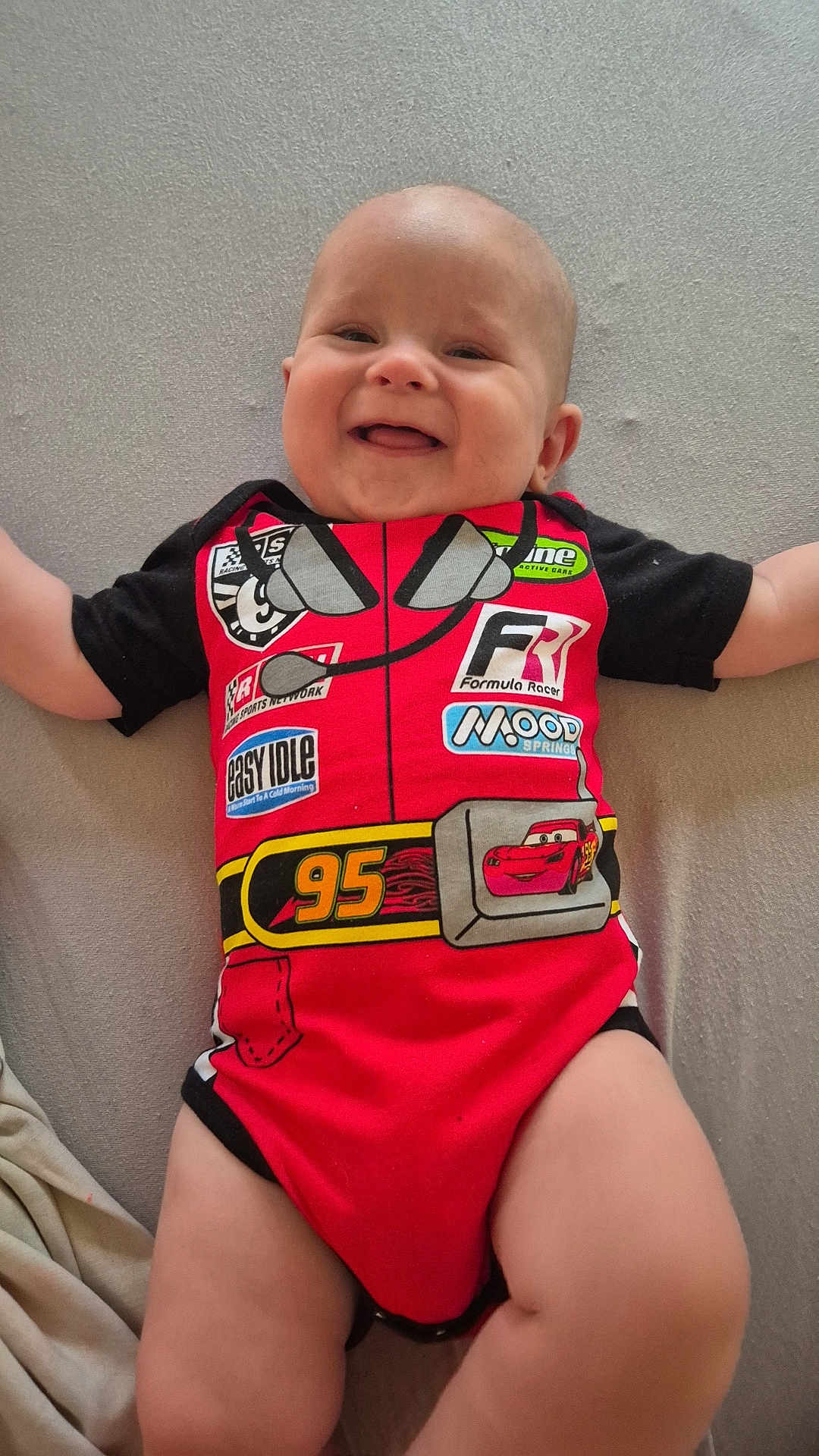 Lyzio a rejoint le concours — aidez-le/la à gagner de superbes lots ! baby, child, smiling, onesie, red_clothing, racing_theme, cartoon, indoor, lying_down, cute, happy, infant, person, skin, head, arms, legs, clothing, fabric, soft_surface