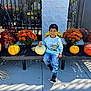 child, pumpkin, bench, flowers, scarecrow, autumn, fall, outdoor, smiling, casual_clothing, sneakers, daylight, plant, leafy_plants, jeans, sitting, decor, holiday, seasonal, portrait