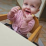 Faith is registered to the contest to win money with this photo: baby, child, high_chair, pink_clothing, smiling, happy, indoor, food, hand, face, person, wooden_chair, tiled_floor, public_space, background_blur, cute, young_child, infant, feeding, seated