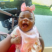 Kaydence is registered to the contest to win money with this photo: baby, child, smiling, pink_bow, bib, dress, hand, stroller, outdoor, fence, grass, concrete, joyful, headband, person, infant, happy, holding, cute, playful