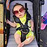Edith is registered to the contest to win money with this photo: baby, child, stroller, sunglasses, neon_clothing, outdoor, portrait, seated, infant, face, fashion, summer, sunlight, pavement, harness, carriage, legs, bare_feet, colorful, playground