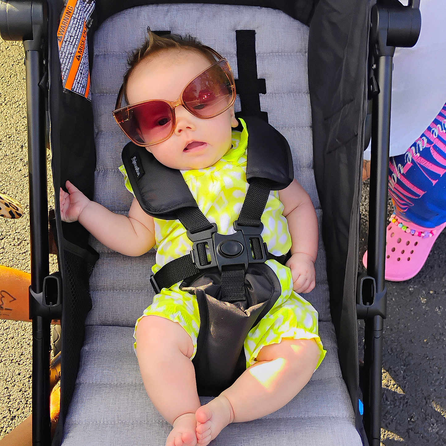 Edith is registered to the contest to win money with this photo: baby, child, stroller, sunglasses, neon_clothing, outdoor, portrait, seated, infant, face, fashion, summer, sunlight, pavement, harness, carriage, legs, bare_feet, colorful, playground