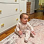 Lucia joined the competition — help win amazing prizes! baby, infant, child, onesie, sitting, rug, floor, dresser, drawer, door, wood_floor, home_interior, curious, patterned_rug, golden_knobs, indoor, cute, small_person, furniture, cozy