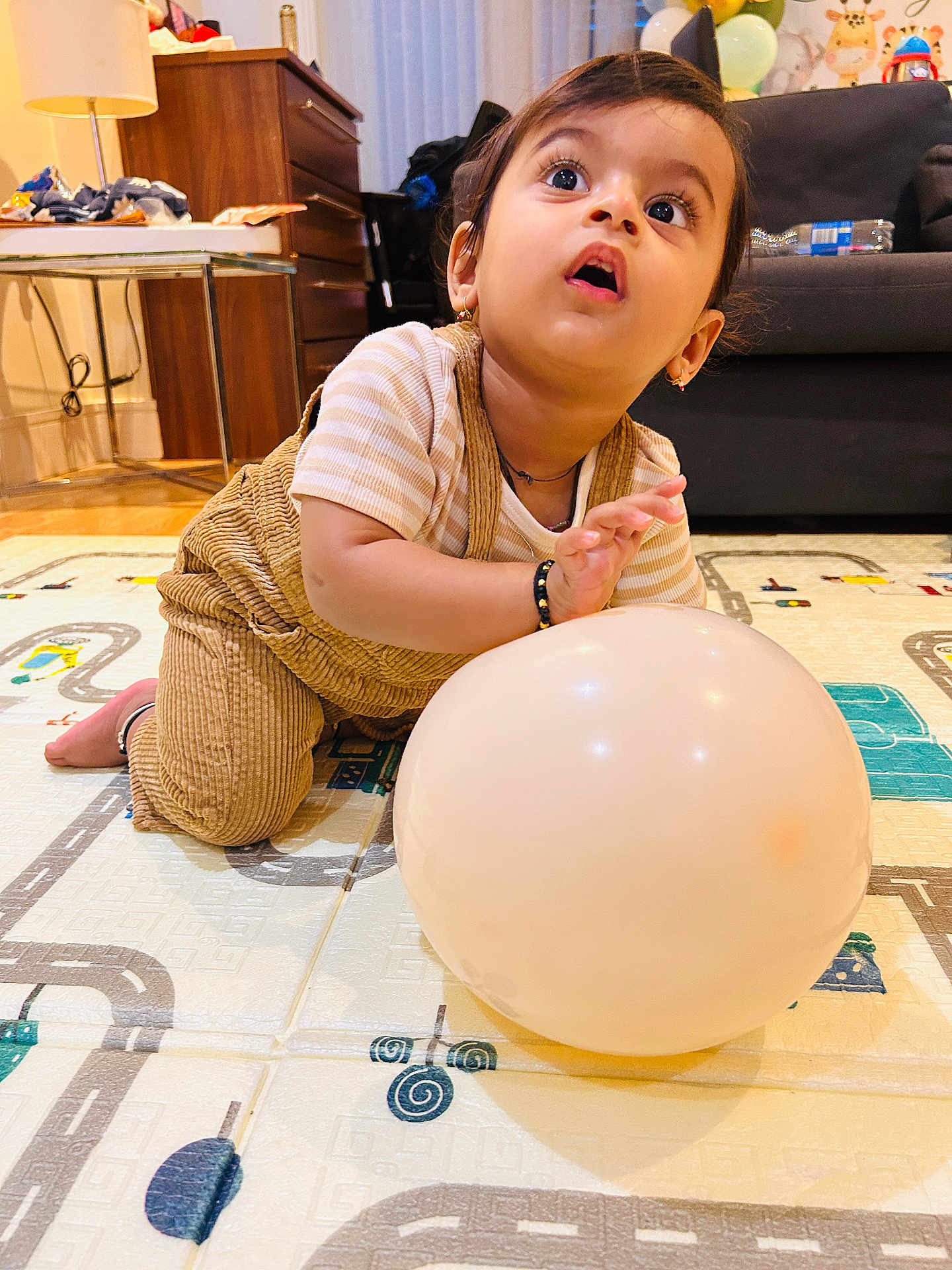 Vihaan is registered to the contest to win money with this photo: toddler, child, balloon, play_mat, indoors, curtains, sofa, lamp, wooden_furniture, bracelet, striped_shirt, overalls, floor_mat, toy, expression, candid, home, cute, curious, earrings