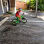 child, bicycle, helmet, training_wheels, outdoor, parking_lot, brick_wall, greenery, pavement, sunlight, shadow, colorful_clothing, red_shirt, brown_pants, sneakers, learning, riding, young, safety_gear, daytime