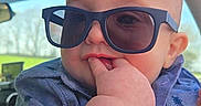 John Wayne is registered to the contest to win money with this photo: baby, child, sunglasses, blue_shirt, hand, face, car_interior, window, outdoor, cute, portrait, person, infant, casual_clothing, skin, head, eyes, fingers, summer, sunlight