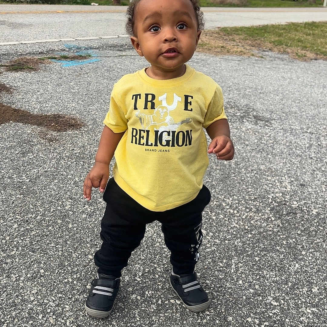Amir Mcintosh is registered to the contest to win money with this photo: toddler, child, standing, yellow_shirt, black_pants, outdoor, pavement, suburban, house, trees, curious, person, footwear, grass, daytime, casual_clothing, sidewalk, young_child, urban, nature