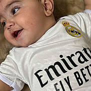 Adam is registered to the contest to win money with this photo: baby, infant, child, smiling, close_up, cheek, eye, ear, real_madrid, soccer_jersey, logo, text_on_clothing, white_shirt, blanket, lying_down, cute, portrait, chubby_cheeks, soft_texture, happy