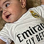baby, infant, child, smiling, close_up, cheek, eye, ear, real_madrid, soccer_jersey, logo, text_on_clothing, white_shirt, blanket, lying_down, cute, portrait, chubby_cheeks, soft_texture, happy