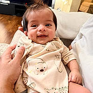 Krishna joined the competition — help win amazing prizes! baby, smile, spiky_hair, onesie, bear_print, crocodile_print, hand, person, indoor, couch, wood_floor, television, happy, infant, cute, child, holding, seated, warm_light, cozy