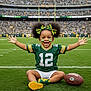 athlete, bows, cheerful, child, crowd, daylight, face_paint, football, football_field, grass_field, green_bay_packers, happy, outdoor, smiling, sports_fan, sports_jersey, stadium, team_spirit, toddler, yellow_shoes