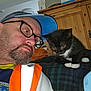 man, glasses, cap, beard, kitten, black_and_white_cat, blanket, plaid, indoor, wooden_furniture, curiosity, pet, animal, portrait, face, close_up, casual_clothing, home, resting, interaction