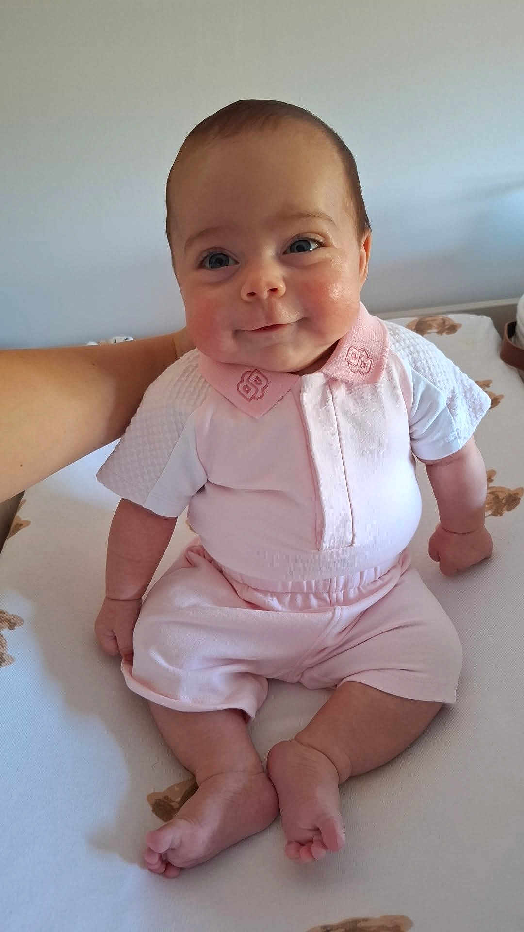 Emy a rejoint le concours — aidez-le/la à gagner de superbes lots ! baby, infant, child, smile, pink_clothing, sitting, cute, chubby_cheeks, bed, adult_arm, soft_lighting, indoors, portrait, happy, short_sleeves, pale_skin, blue_eyes, casual, home, cozy