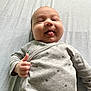 baby, infant, tongue_out, thumbs_up, grey_clothing, stars_pattern, bed, bedding, smile, child, cute, happy, person, portrait, laying_down, soft_texture, casual, indoors, playful, young