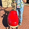 animal, brick_pavement, casual_clothing, cat, daylight, footwear, hawaiian_shirt, jeans, leash, outdoor, person, pet, playful, red_shoe, sculpture, shadow, standing, street_art, sunny_day, tabby_cat