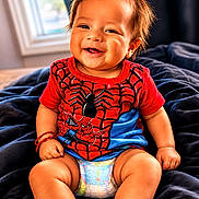 Enzo is registered to the contest to win money with this photo: baby, smiling, child, diaper, red_shirt, spiderman, indoor, blanket, happy, cute, toddler, sitting, hair, face, person, portrait, skin, clothing, window, bracelet