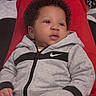 baby, child, curly_hair, grey_tracksuit, nike, hoodie, casual_wear, infant, portrait, indoors, red_background, relaxed, cute, hand, face, fashion, young, sitting, soft_lighting, expression