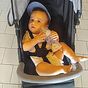 Loucas participe au concours pour gagner de l'argent avec cette photo : baby, baby_bottle, baby_girl_or_boy, black_stroller, blue_cap, bottle, casual_clothing, child, curious_expression, cute, footwear_absent, holding, indoors, infant, light_skin, seatbelt, sitting, stroller, tiled_floor, young_child