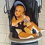 baby, baby_bottle, baby_girl_or_boy, black_stroller, blue_cap, bottle, casual_clothing, child, curious_expression, cute, footwear_absent, holding, indoors, infant, light_skin, seatbelt, sitting, stroller, tiled_floor, young_child