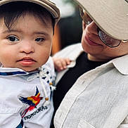 Ethan is registered to the contest to win money with this photo: baby, child, adult, portrait, close_up, face, smiling, serious_expression, hat, cap, sunglasses, outdoor, cuddling, family, parent, clothing, white_shirt, embroidered_bird, photography, blurred_background