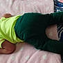 baby, infant, child, blanket, bed, bedding, clothing, yellow_shirt, green_pants, lying_down, soft_texture, patterned_bedding, indoors, person, head, face, arm, legs, feet, curious_expression
