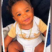 Jamari is registered to the contest to win money with this photo: baby, child, infant, smiling, portrait, sitting, curly_hair, dark_skin, onesie, clothing, necklace, bracelet, socks, blanket, cushion, heart, toy_blocks, teddy_bear, indoor, happy