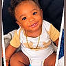 baby, child, infant, smiling, portrait, sitting, curly_hair, dark_skin, onesie, clothing, necklace, bracelet, socks, blanket, cushion, heart, toy_blocks, teddy_bear, indoor, happy