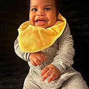 Shanan joined the competition — help win amazing prizes! baby, child, smiling, yellow_bib, grey_clothing, portrait, happy, cute, infant, sitting, hands, face, curly_hair, indoor, closeup, young_child, expression, bright, warm, adorable